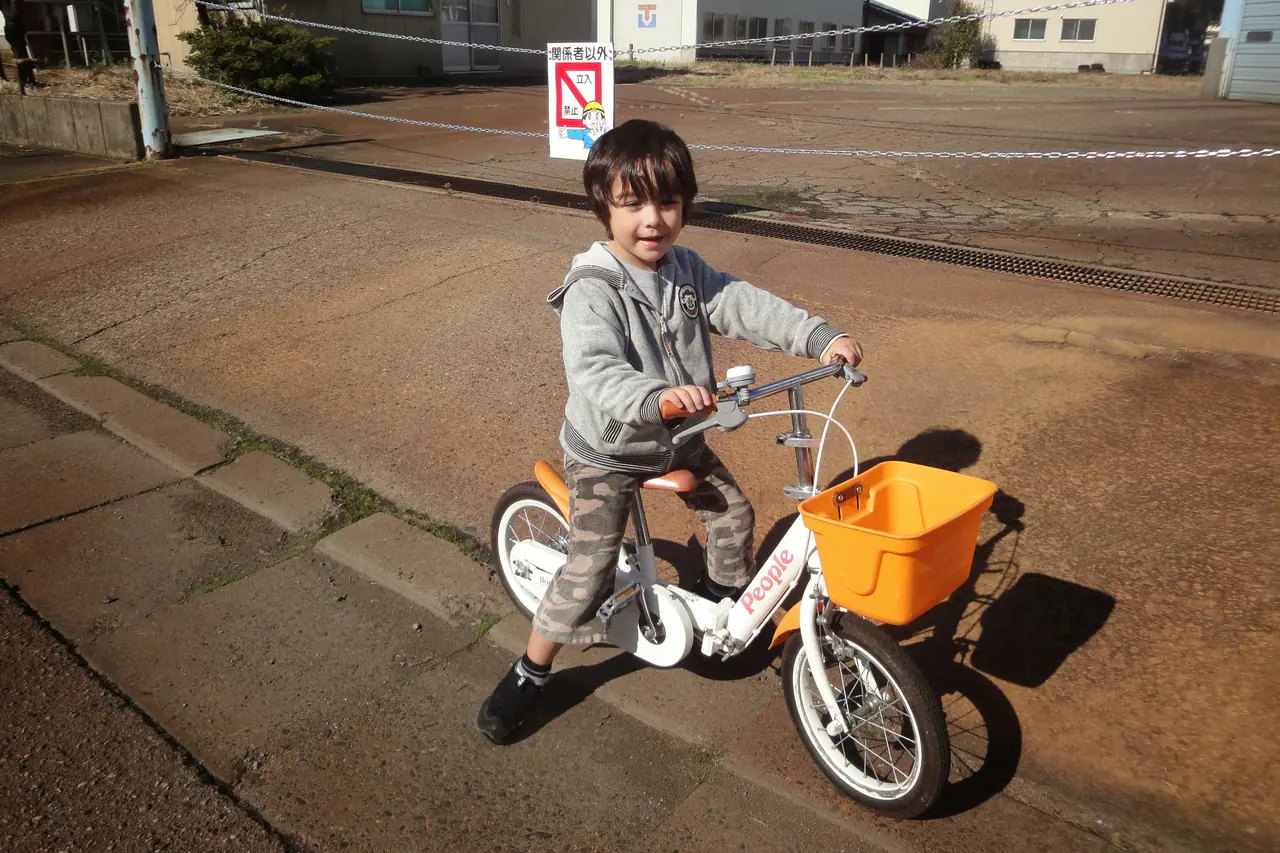 A young boy on a small white bicycle with an orange basket, on a quiet street in Japan