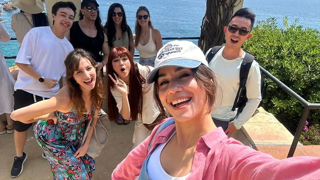 A group selfie of the Dreaming French team on a coastal path above the sea, everyone laughing at the camera